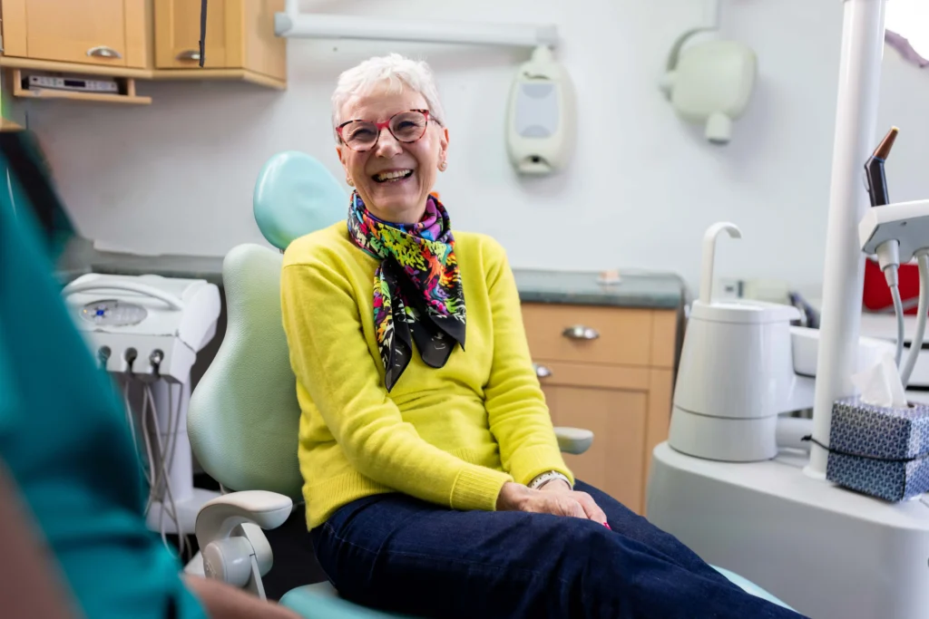 A cheerful senior woman wearing a bright yellow sweater and colourful scarf, sitting comfortably in a dental chair.